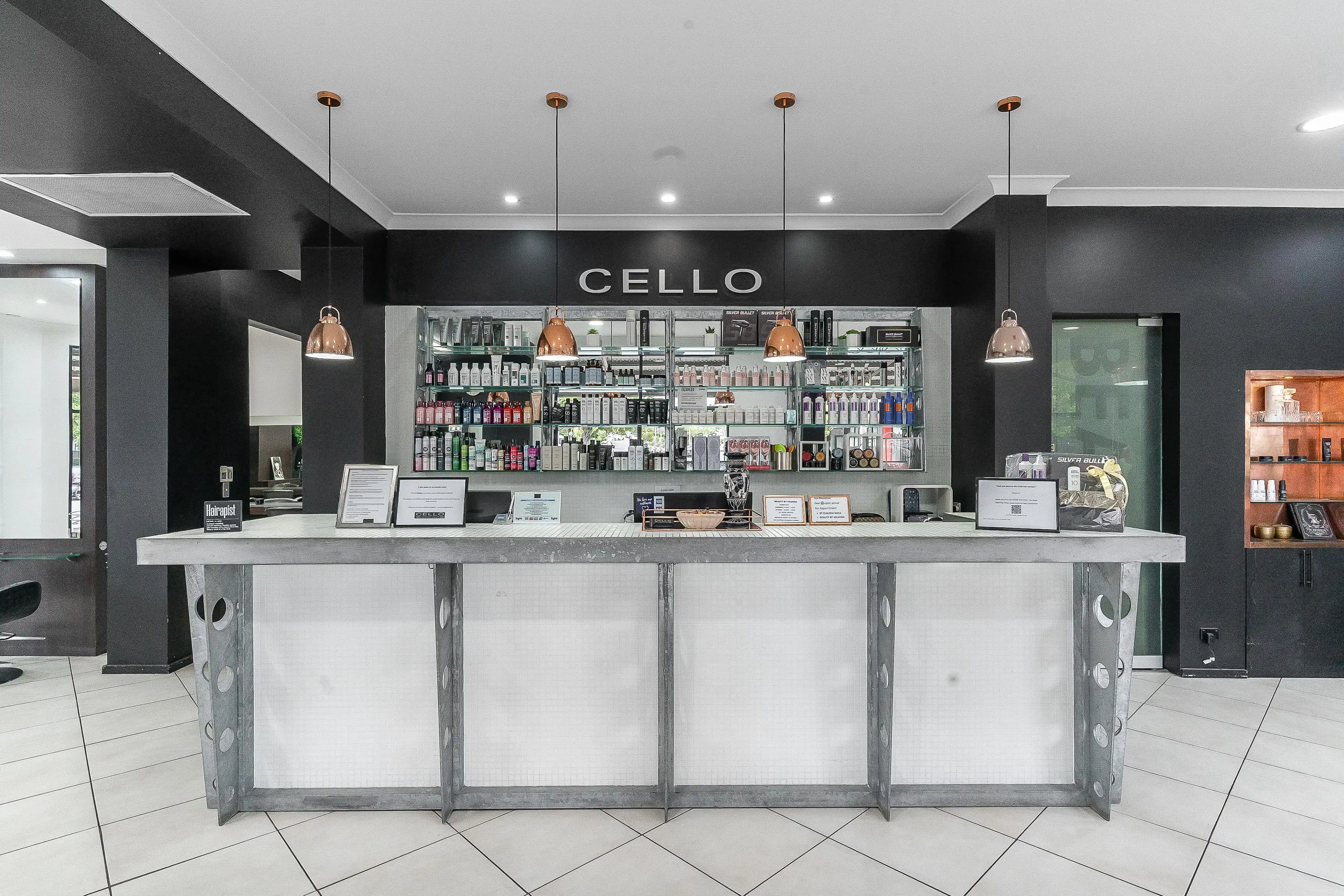 The reception desk at Cello Hairdressers, with copper pendant lights and the lit product wall under the CELLO sign.