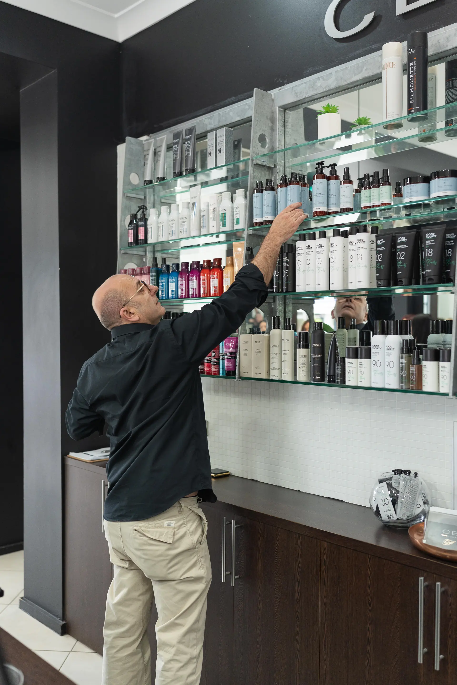 Reaching for a product on the glass shelves of the retail wall at Cello Hairdressers.