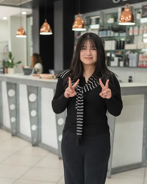 Mickey, salon assistant at Cello Hairdressers, smiling with both hands raised in peace signs.