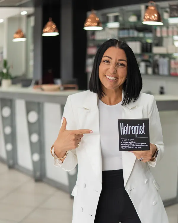 Tanya, senior stylist at Cello Hairdressers, smiling and pointing to a Hairapist sign.
